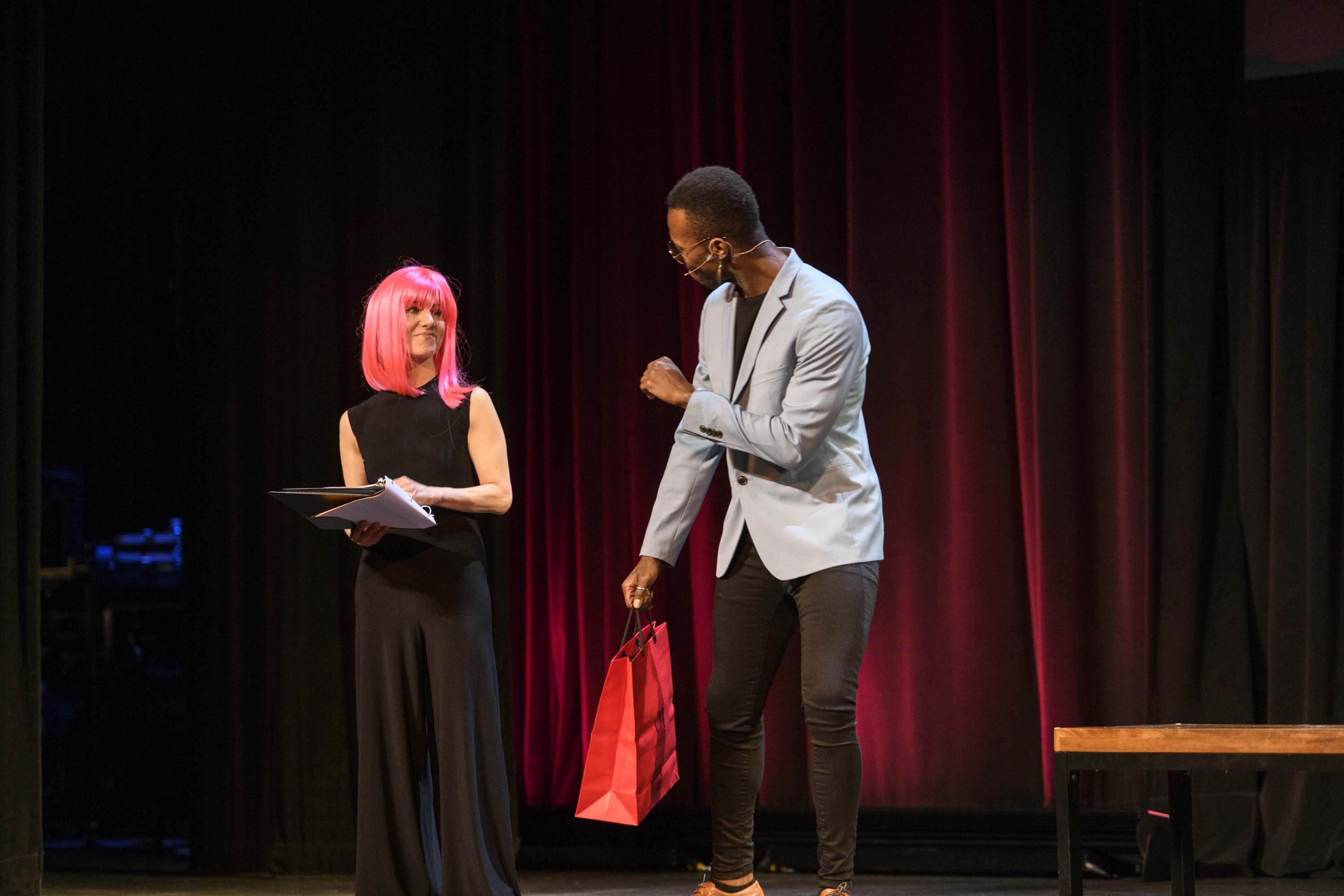Woman in pink wig talks to man on stage