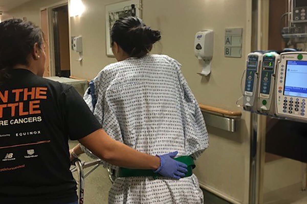 Female patient with back to camera being helped down a hospital hallway.