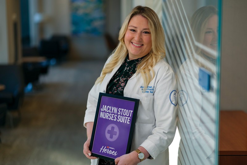 Jakki Stout stands in her white coat and holds a sign for the nurses’ suite that was named after her.