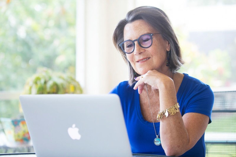 A woman with short greyish-brown hair wearing glasses and a blue shirt smiles while looking at a laptop. She rests her chin on her hand, appearing engaged and thoughtful. In the background, there are soft-focus green plants and bright natural light, creating a warm and inviting atmosphere.