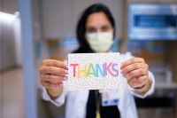 A masked nurse holds a card that says “thanks” in bright colors